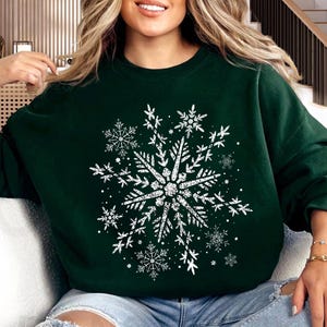 May include: Dark green sweatshirt featuring a large, distressed white snowflake design. The snowflake is the focal point, surrounded by smaller snowflake accents. The sweatshirt is a comfortable, casual style, perfect for winter or holiday wear.