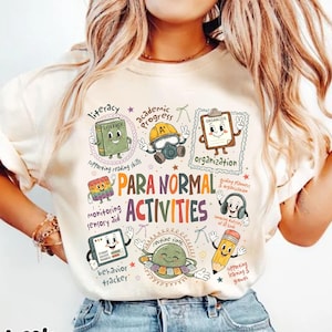 May include: An ivory t-shirt featuring a colorful graphic with the text "PARA NORMAL ACTIVITIES" surrounded by cartoon illustrations of school supplies including a book, clipboard, pencil, and sensory toy, each labeled with educational terms.