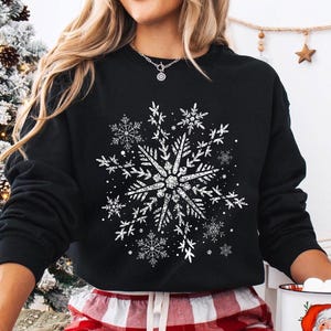 May include: Black sweatshirt featuring a large, detailed white snowflake design. The snowflake is the focal point, surrounded by smaller snowflakes. The person is wearing red and white plaid pajama pants. A mug with a Santa Claus illustration is held in the hand.