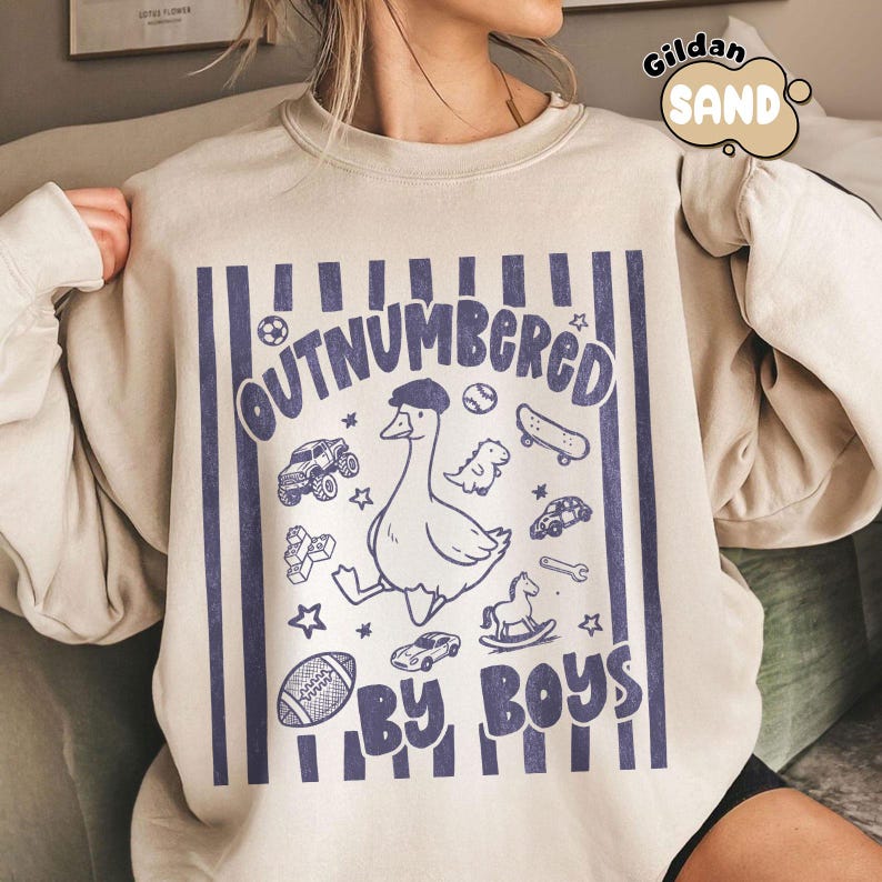 May include: Sand-colored sweatshirt with a blue graphic that reads "OUTNUMBERED BY BOYS." The graphic features a goose wearing a hat, surrounded by toy cars, a skateboard, and a football. The sweatshirt has a crew neck and long sleeves.