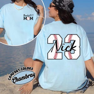 Może przedstawiać: Jasnoniebieska koszulka z motywem baseballowym i napisem "Baseball Mom" z przodu oraz numerem "23" z imieniem "Nick" z tyłu.