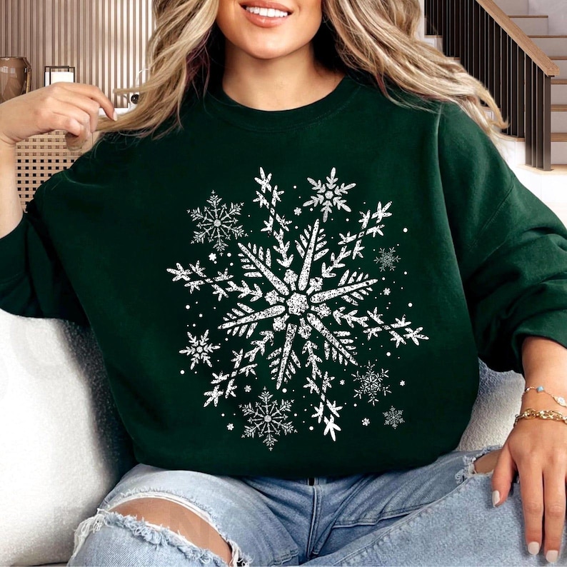 May include: Dark green sweatshirt featuring a large, distressed white snowflake design. The snowflake is the focal point, surrounded by smaller snowflake accents. The sweatshirt is a comfortable, casual style, perfect for winter or holiday wear.