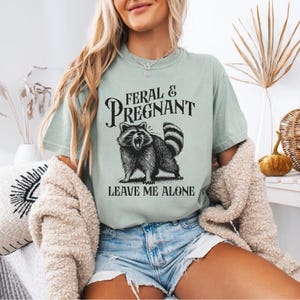 K&ouml;nnte beinhalten: Hellgr&uuml;nes T-Shirt mit dem Text "FERAL & PREGNANT" &uuml;ber einer schwarz-wei&szlig;en Illustration eines Waschb&auml;ren. Darunter steht "LEAVE ME ALONE". Das Shirt besteht aus einem weichen Material.