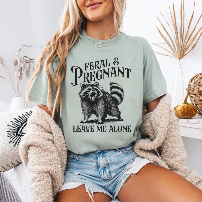K&ouml;nnte beinhalten: Hellgr&uuml;nes T-Shirt mit dem Text "FERAL & PREGNANT" &uuml;ber einer schwarz-wei&szlig;en Illustration eines Waschb&auml;ren. Darunter steht "LEAVE ME ALONE". Das Shirt besteht aus einem weichen Material.