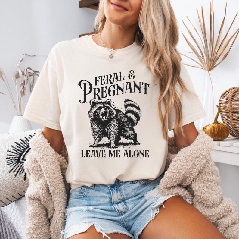 May include: A white t-shirt with a black and white illustration of a raccoon. The text on the shirt reads 'Feral & Pregnant Leave Me Alone'. The shirt is being worn by a person.