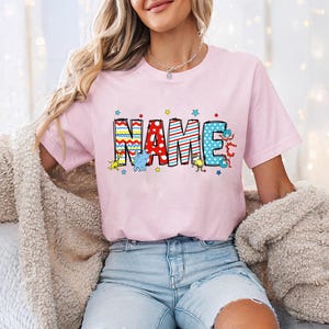 May include: Pink t-shirt with a colorful "NAME" graphic featuring a whimsical design with stars and characters. The letters are made of different patterns and colors, including red, blue, yellow, and white.