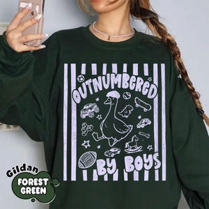 May include: Forest green sweatshirt with the text "OUTNUMBERED BY BOYS" in white. The design includes a cartoon goose wearing a hat, along with illustrations of a football, cars, a skateboard, and other toys. The brand name "Gildan" is also visible.