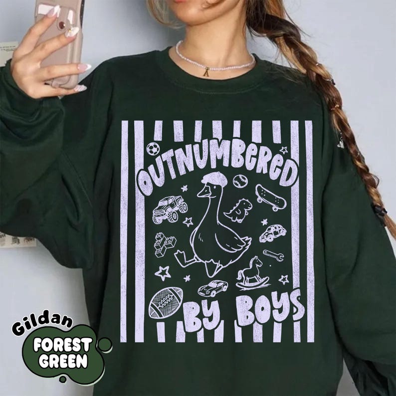 May include: Forest green sweatshirt with the text "OUTNUMBERED BY BOYS" in white. The design includes a cartoon goose wearing a hat, along with illustrations of a football, cars, a skateboard, and other toys. The brand name "Gildan" is also visible.
