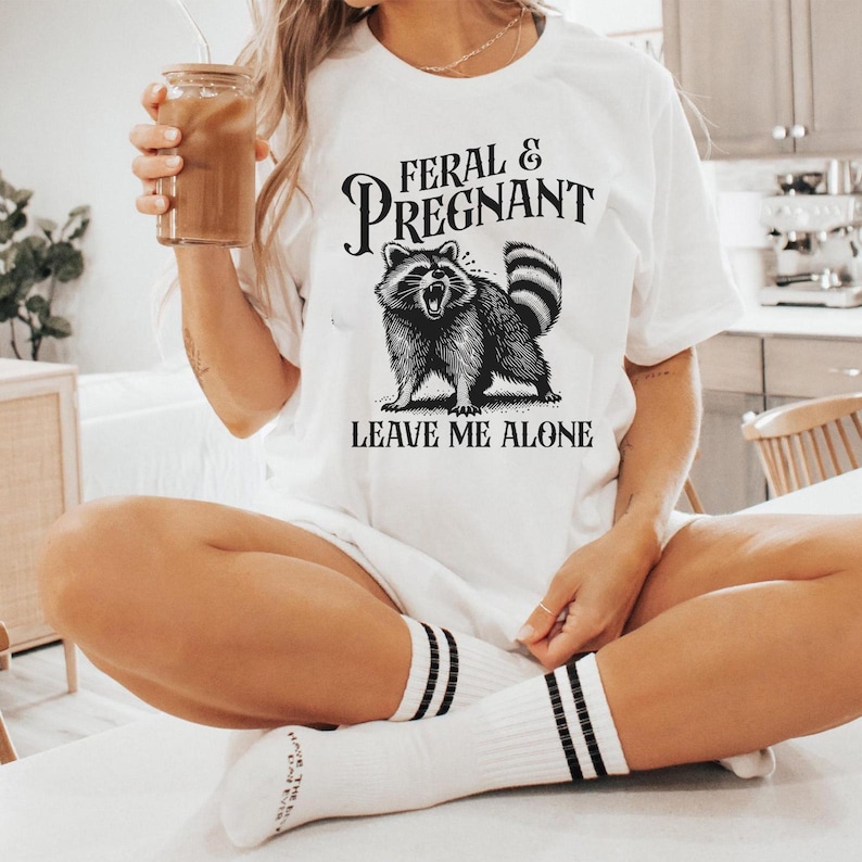 K&ouml;nnte beinhalten: Wei&szlig;es T-Shirt mit dem Text "FERAL & PREGNANT" &uuml;ber einer schwarz-wei&szlig;en Illustration eines Waschb&auml;ren. Der Text "LEAVE ME ALONE" steht unter dem Waschb&auml;ren. Das Shirt wird mit wei&szlig;en Socken mit schwarzen Streifen getragen.