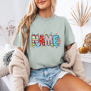 May include: A light green t-shirt with the word "NAME" in colorful, patterned letters. The letters are decorated with stars and cartoon characters.