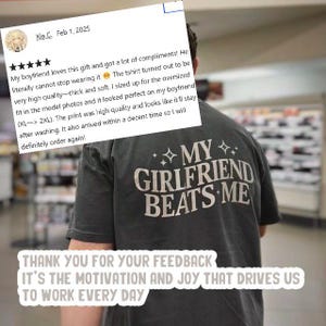May include: A dark gray t-shirt with the text "MY GIRLFRIEND BEATS ME" printed in white. The t-shirt is being worn by a person in a store. The image also includes a customer review with a five-star rating and a positive comment about the t-shirt.