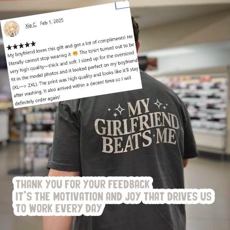 May include: A dark gray t-shirt with the text "MY GIRLFRIEND BEATS ME" printed in white. The t-shirt is being worn by a person in a store. The image also includes a customer review with a five-star rating and a positive comment about the t-shirt.
