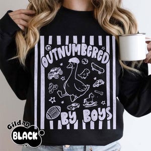 May include: Black sweatshirt with the text "OUTNUMBERED BY BOYS" in white. The design features a cartoon goose wearing a hat, surrounded by toy cars, a football, a skateboard, and other toys. The brand name "Gildan BLACK" is printed on the bottom left.