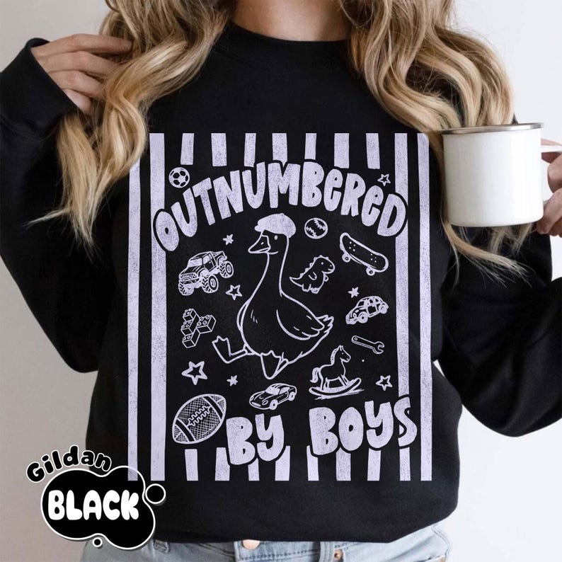 May include: Black sweatshirt with the text "OUTNUMBERED BY BOYS" in white. The design features a cartoon goose wearing a hat, surrounded by toy cars, a football, a skateboard, and other toys. The brand name "Gildan BLACK" is printed on the bottom left.