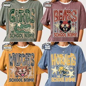 May include: Four short-sleeve t-shirts in moss, espresso, mustard, and blue jean colors. Each shirt features a graphic with a mascot, stars, vertical stripes, and the text SCHOOL NAME. Mascots include a gator, bear, wildcat, and hornet.