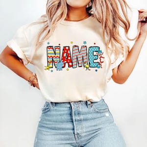 May include: A white t-shirt with the word "NAME" printed in colorful letters. The letters are decorated with stripes, polka dots, and zigzags. There are small cartoon characters around the letters.