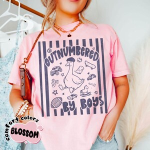 May include: Pink t-shirt with a graphic that reads "OUTNUMBERED BY BOYS." The design features a cartoon goose wearing a hat, surrounded by toy cars, a skateboard, and a football. The shirt has vertical navy blue stripes.