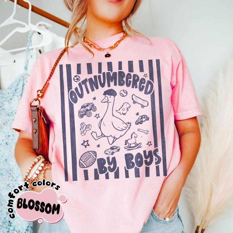 May include: Pink t-shirt with a graphic that reads "OUTNUMBERED BY BOYS." The design features a cartoon goose wearing a hat, surrounded by toy cars, a skateboard, and a football. The shirt has vertical navy blue stripes.