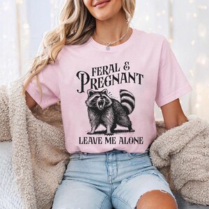 K&ouml;nnte beinhalten: Hellrosa T-Shirt mit dem Text "FERAL & PREGNANT" &uuml;ber einer schwarz-wei&szlig;en Illustration eines Waschb&auml;ren. Darunter steht der Text "LEAVE ME ALONE".