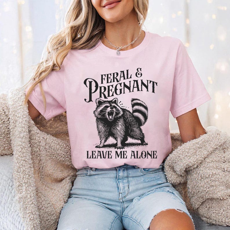 K&ouml;nnte beinhalten: Hellrosa T-Shirt mit dem Text "FERAL & PREGNANT" &uuml;ber einer schwarz-wei&szlig;en Illustration eines Waschb&auml;ren. Darunter steht der Text "LEAVE ME ALONE".