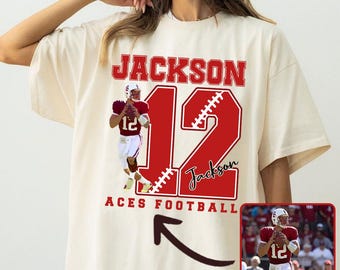 May include: Cream-colored t-shirt with a red football-themed design. The design includes the name "JACKSON" above a large number "12" with football stitching, a football player graphic, and the words "ACES FOOTBALL".