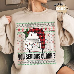 May include: Beige sweatshirt with a festive design. The graphic features a red and black plaid hat with white fur trim, adorned with Christmas lights and holly. The text "YOU SERIOUS CLARK?" is printed below the hat.