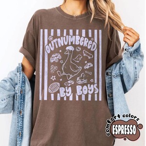 May include: Espresso-colored t-shirt with the text "OUTNUMBERED BY BOYS" in a light purple font. The design includes a cartoon goose, toy cars, a skateboard, and a football. The shirt has a vintage, worn look.