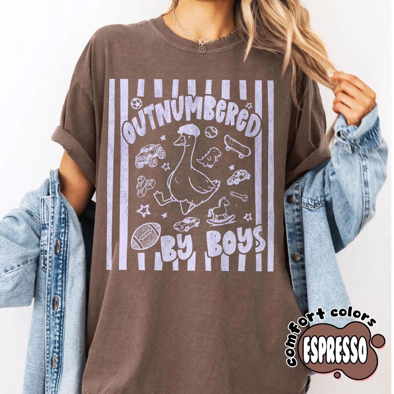 May include: Espresso-colored t-shirt with the text "OUTNUMBERED BY BOYS" in a light purple font. The design includes a cartoon goose, toy cars, a skateboard, and a football. The shirt has a vintage, worn look.
