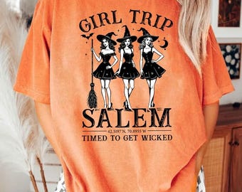 May include: An orange short-sleeve t-shirt featuring a black graphic of three witches with the text GIRL TRIP SALEM 42.5197 N. 70.8955 W. TIMED TO GET WICKED. The design includes a broom, a bat, and a crescent moon, printed in a vintage style.