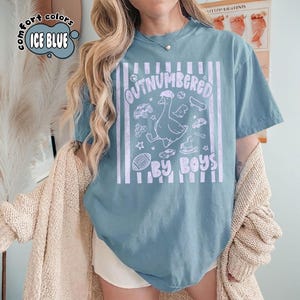 May include: Ice blue t-shirt with the text "OUTNUMBERED BY BOYS" and a graphic of a goose surrounded by toys. The shirt has a vintage, distressed look and vertical white stripes.