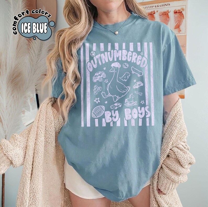 May include: Ice blue t-shirt with the text "OUTNUMBERED BY BOYS" and a graphic of a goose surrounded by toys. The shirt has a vintage, distressed look and vertical white stripes.