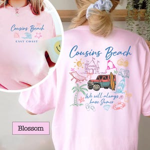 Op de afbeelding: Lichtroze T-shirt met de woorden "Cousins Beach" in blauw schrift. De achterkant van het shirt heeft een kleurrijke strandthema-afbeelding met een jeep, surfboard en andere zomerelementen. De tekst "We will always have Summer" is ook opgenomen.