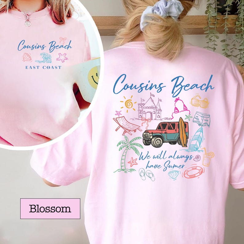 Op de afbeelding: Lichtroze T-shirt met de woorden "Cousins Beach" in blauw schrift. De achterkant van het shirt heeft een kleurrijke strandthema-afbeelding met een jeep, surfboard en andere zomerelementen. De tekst "We will always have Summer" is ook opgenomen.