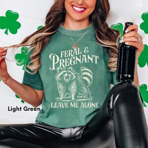 K&ouml;nnte beinhalten: Hellgr&uuml;nes T-Shirt mit dem Text "FERAL & PREGNANT" &uuml;ber einer Waschb&auml;r-Illustration und den Worten "LEAVE ME ALONE" darunter. Das Shirt hat eine hellgr&uuml;ne Farbe.