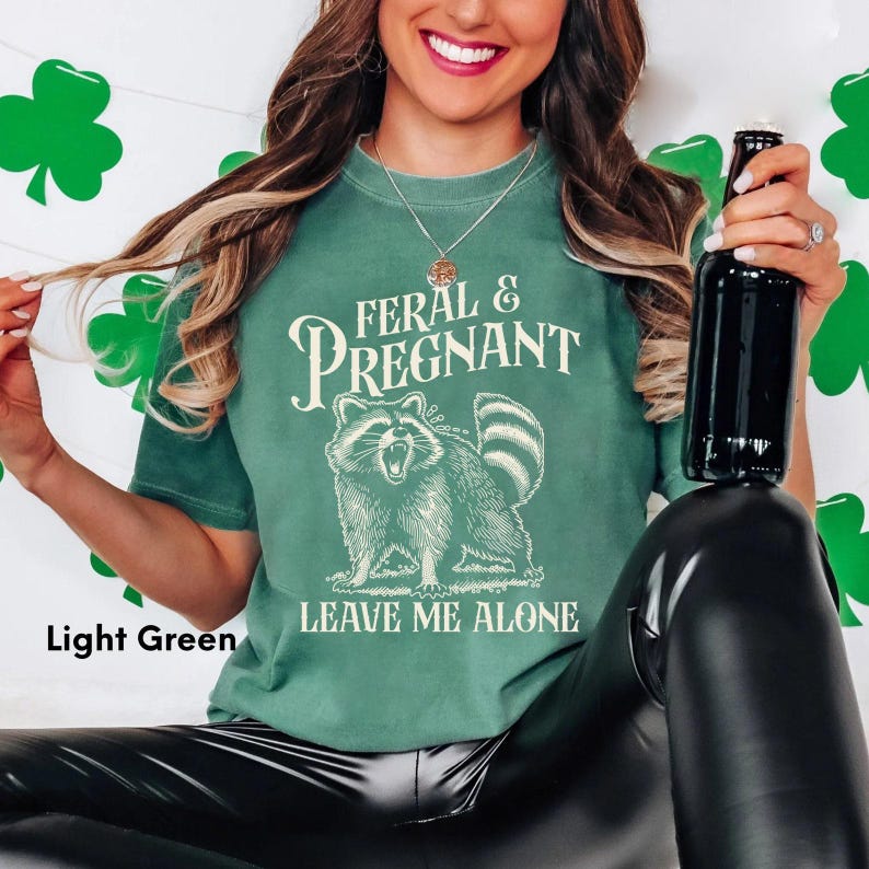 K&ouml;nnte beinhalten: Hellgr&uuml;nes T-Shirt mit dem Text "FERAL & PREGNANT" &uuml;ber einer Waschb&auml;r-Illustration und den Worten "LEAVE ME ALONE" darunter. Das Shirt hat eine hellgr&uuml;ne Farbe.