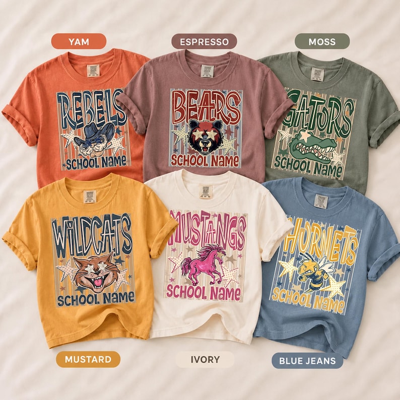 May include: Six short-sleeve t-shirts in yam, espresso, moss, mustard, ivory, and blue jeans colors. Each shirt features a graphic with a mascot, stars, vertical stripes, and the text SCHOOL NAME. Mascots include a cowboy, bear, alligator, cat, horse, and hornet.