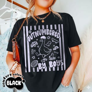 May include: Black t-shirt with a white graphic that reads "OUTNUMBERED BY BOYS." The graphic features a cartoon goose wearing a hat, surrounded by toy cars, a football, a skateboard, and other toys. The words "comfort colors" and "BLACK" are also printed on the shirt.