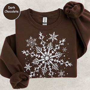 May include: Dark chocolate-colored sweatshirt with a large white snowflake design. The snowflake is composed of intricate details and surrounded by smaller snowflakes. The sweatshirt has a crew neck and long sleeves. The text "Dark Chocolate" is in a circle in the upper left corner.