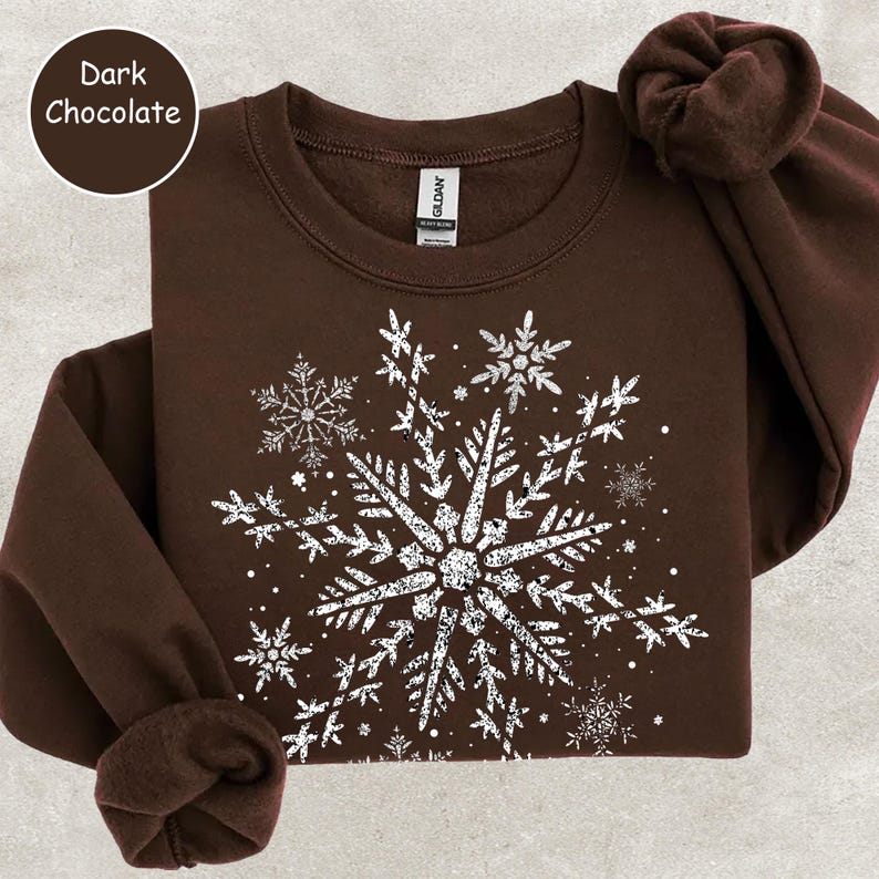 May include: Dark chocolate-colored sweatshirt with a large white snowflake design. The snowflake is composed of intricate details and surrounded by smaller snowflakes. The sweatshirt has a crew neck and long sleeves. The text "Dark Chocolate" is in a circle in the upper left corner.