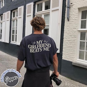 May include: A person wearing a black t-shirt with the text "My Girlfriend Beats Me" printed on the back. The person is standing in front of a building with white windows.
