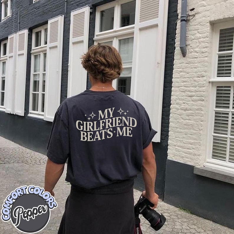 May include: A person wearing a black t-shirt with the text "My Girlfriend Beats Me" printed on the back. The person is standing in front of a building with white windows.