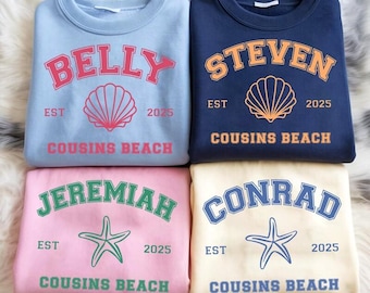 Peut inclure: Quatre sweat-shirts à col rond, bleu clair, marine, rose et crème. Chacun présente un nom en lettres arquées, un coquillage ou une étoile de mer, le texte "EST 2025" et "COUSINS BEACH". Les sweat-shirts sont présentés sur une surface blanche et moelleuse.
