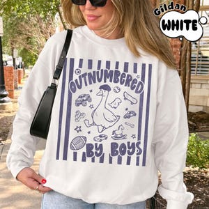 May include: White sweatshirt with a blue graphic that reads "OUTNUMBERED BY BOYS." The graphic features a goose wearing a hat, surrounded by toy cars, a skateboard, and a football. The sweatshirt has a crew neck and long sleeves.
