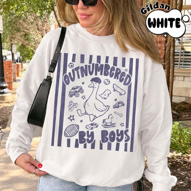 May include: White sweatshirt with a blue graphic that reads "OUTNUMBERED BY BOYS." The graphic features a goose wearing a hat, surrounded by toy cars, a skateboard, and a football. The sweatshirt has a crew neck and long sleeves.