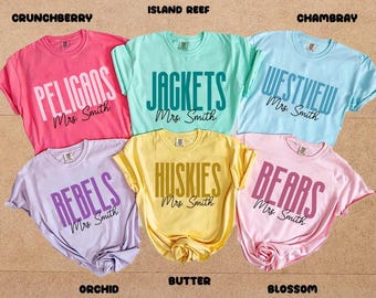 May include: Six short-sleeved t-shirts in various colors, including pink, teal, and yellow. Each shirt features a team name in bold letters, such as "Pelicans" and "Jackets", with "Mrs. Smith" below. The shirts are arranged on a light brown surface.