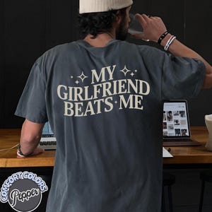 May include: A dark gray t-shirt with the text "MY GIRLFRIEND BEATS ME" in white lettering with small stars on either side of the text.