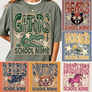 May include: A sage green t-shirt featuring a graphic of a green alligator and the text GATORS SCHOOL NAME. Additional designs include a bear, hornet, cowboy, wildcat, and horse with text like BEARS, HORNETS, REBELS, WILDCATS, and MUSTANGS.