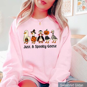 May include: Pink sweatshirt featuring a graphic of five cartoon geese dressed in Halloween costumes: a ghost, witch, vampire, pumpkin, and skeleton. The text "Just A Spooky Goose" is printed below the geese.