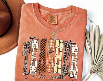Puede incluir: Una camiseta Comfort Colors de color coral con la palabra "TEACHER" en una fuente decorativa, con patrones como estampado de leopardo, calabazas y murciélagos. El nombre "Mrs. Smith" está impreso debajo. Un collar dorado está drapeado sobre la camisa.