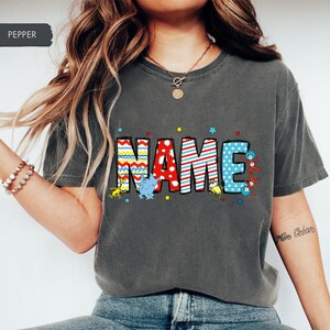 May include: A gray t-shirt with the word "NAME" printed in colorful letters. The letters are decorated with stripes, polka dots, and other patterns. The word "NAME" is surrounded by small stars and cartoon characters.
